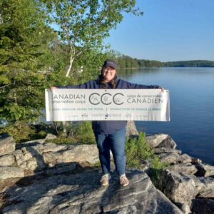 Melanie Beaulieu holding a Canadian Conservation Corps banner on the shoreline of a body of water.