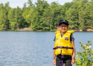 Child wearing a Kawartha Land Trust hat and a lifejacket standing beside Stoney Lake.